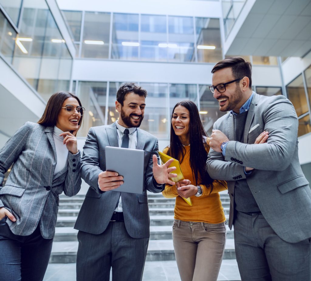 Four successful smiling businesspeople standing in front of corporate building and looking at tablet that man in the middle holding.